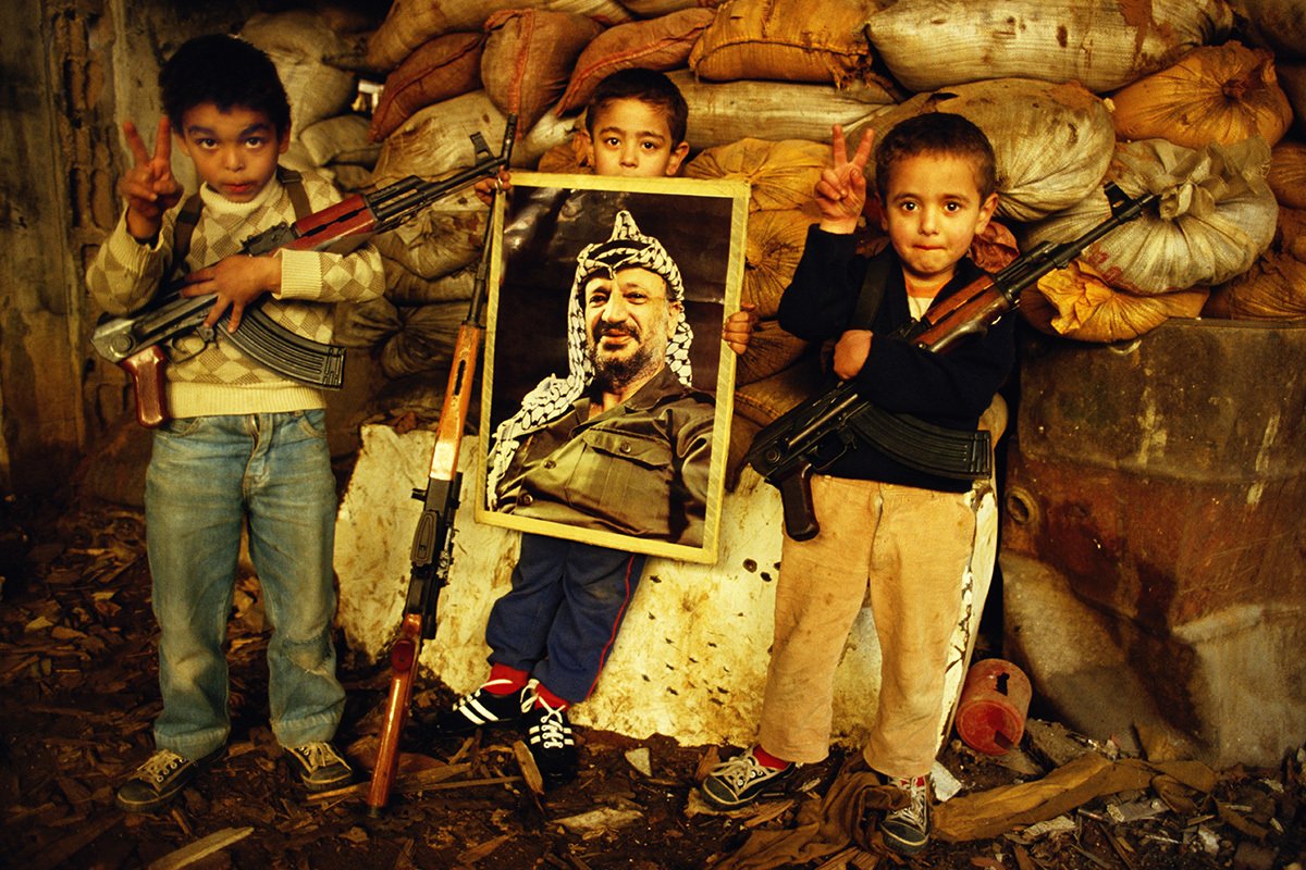 Маленькие палестинские мальчики с автоматами АК-47 и плакатом с изображением президента Ясира Арафата подают знак «V» за победу в лагере беженцев Бурдж аль-Бараджнех в Бейруте, апрель 1987 года. Фото: Tom Stoddart / Getty Images